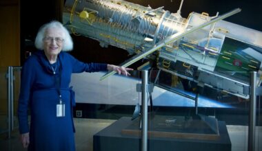 an elderly white-haired woman in a blue dress stands next to a model of a silver space telescope