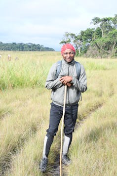 A man stands in grasslands with a hiking stick and a small backpack on