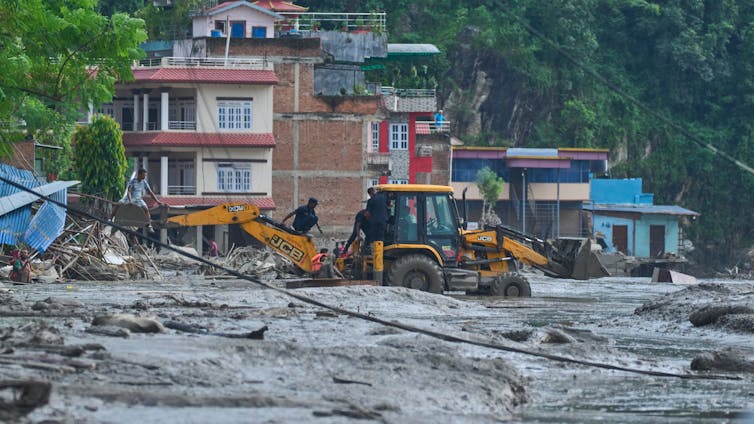 A large piece of equipment like a bulldozer digs in a large expanse of mud with houses in the background.