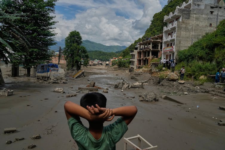 A boy photographed from behind with his hands behind his head looks at a vast expanse of mud and rubble.