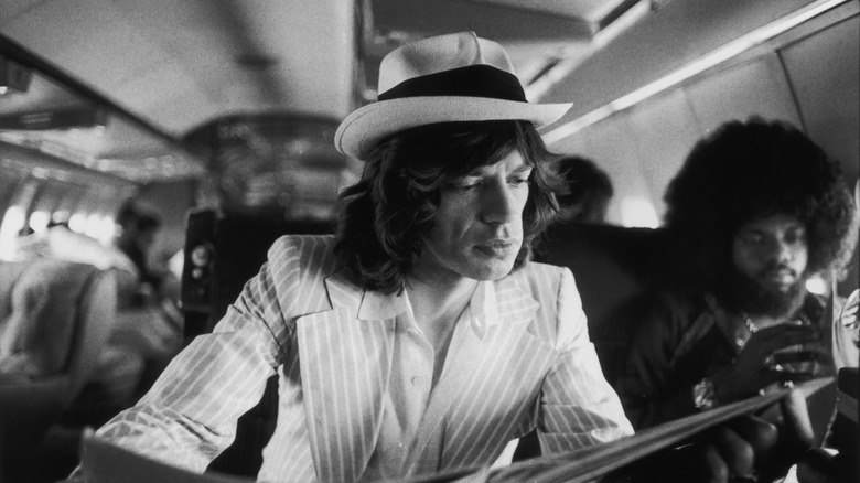 A black-and-white photo of Mick Jagger in a striped suit, reading on an airplane, in 1975
