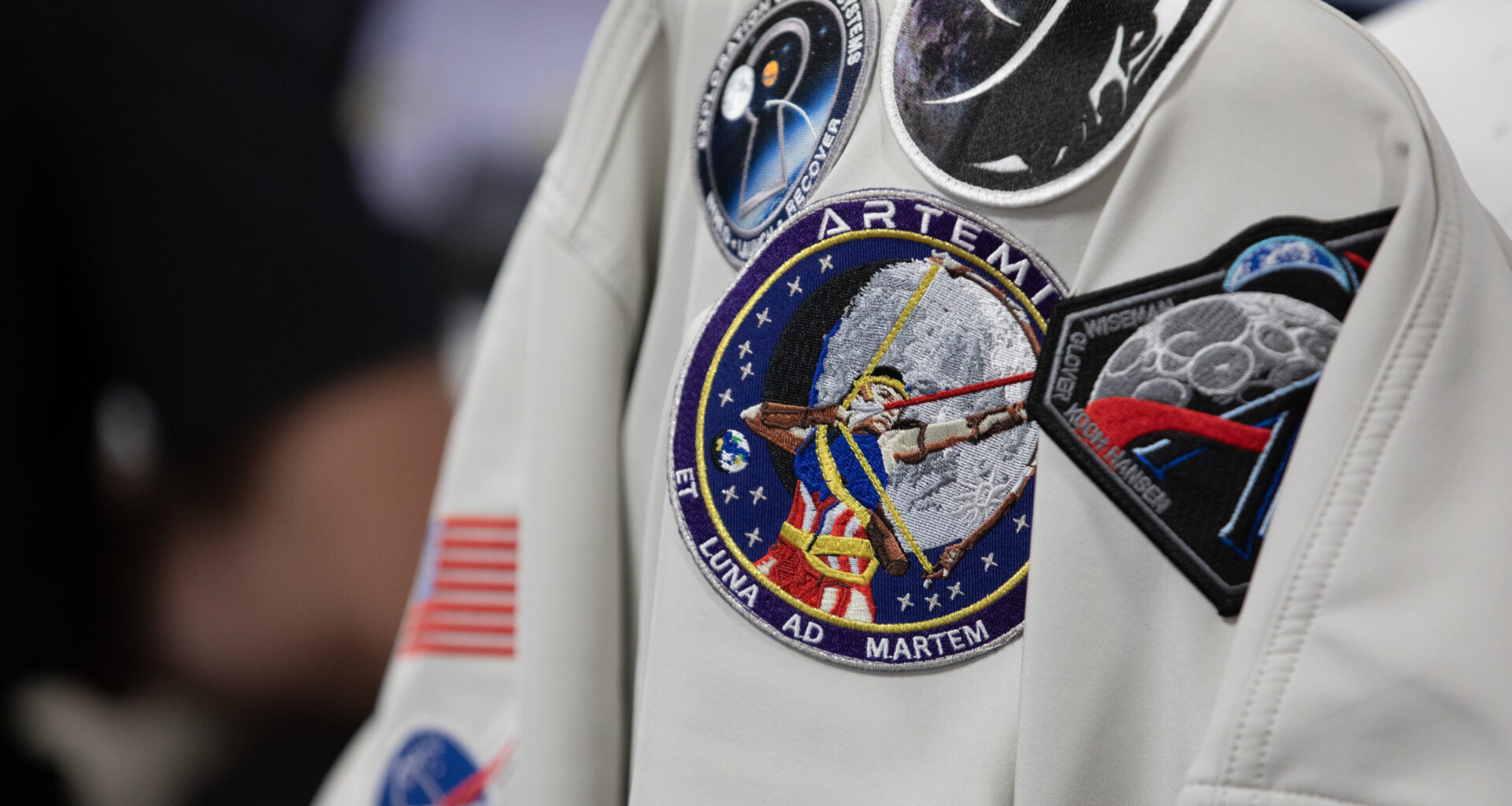 A gray colored jacket with five Artemis mission patches and a NASA insignia hangs on a chair.