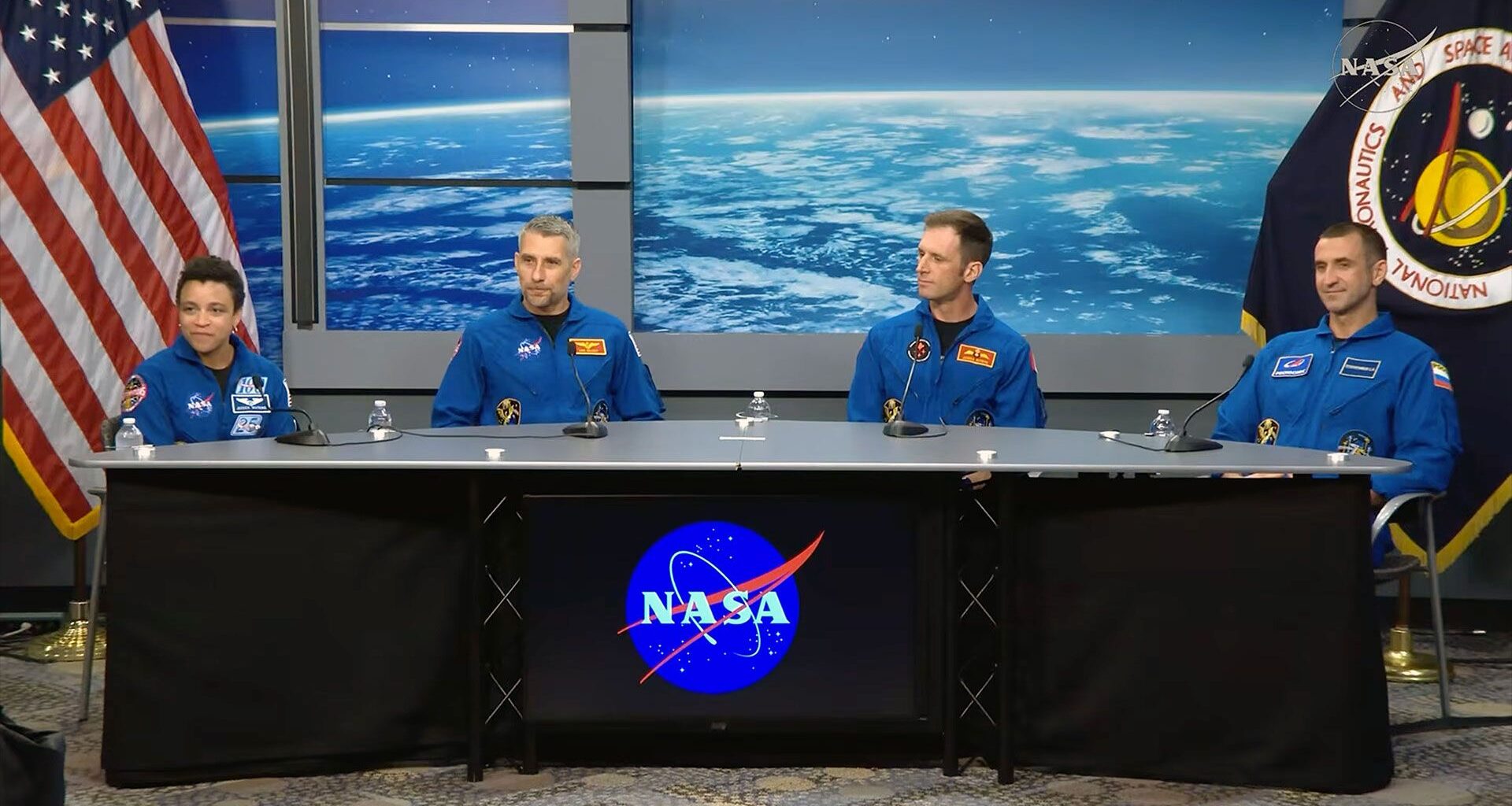 a woman and three men wearing blue coveralls sit at a table with the NASA logo on its front and flags to both sides.