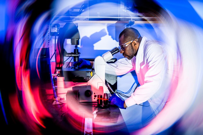 Emeka Okeke looks through a microscope in a lab, wearing goggles and a white lab coat, with a circular blur of red and blue light surrounding him in the frame.