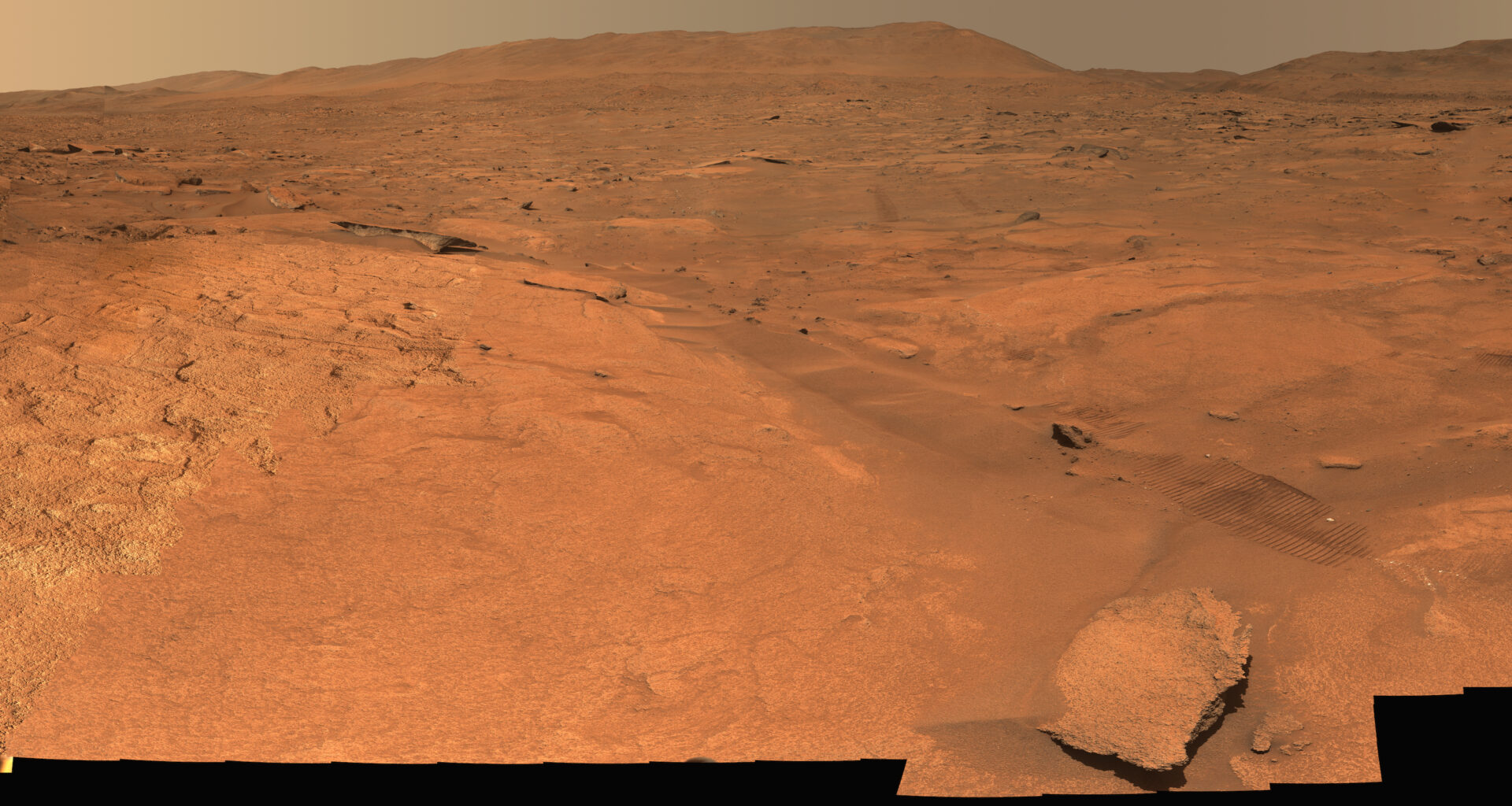A panoramic view of a barren, reddish-orange Martian landscape stretching toward distant hills, partially obscured by the jagged black silhouettes of a rover in the foreground.