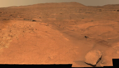 A panoramic view of a barren, reddish-orange Martian landscape stretching toward distant hills, partially obscured by the jagged black silhouettes of a rover in the foreground.