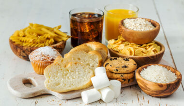 a selection of carbohydrates on a table: sliced bread, pasta, marshmellows, soda and juice, rice, potato chips, chocolate chip cookies and oats