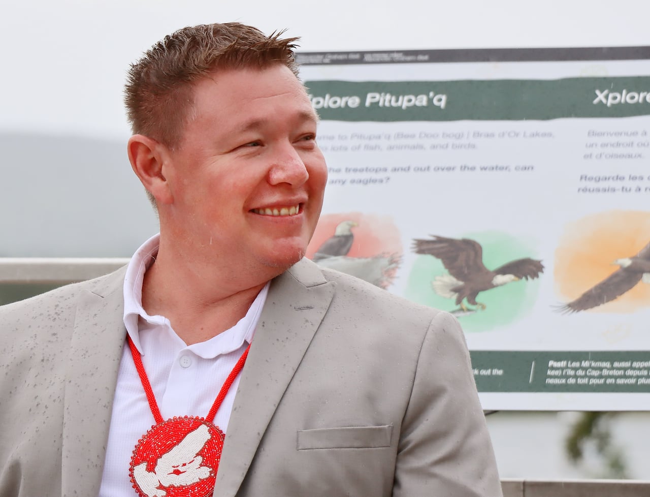 A man with short brown hair wearing a grey suit jacket and white shirt, with a red medallion hanging from his neck, smiles and looks away.