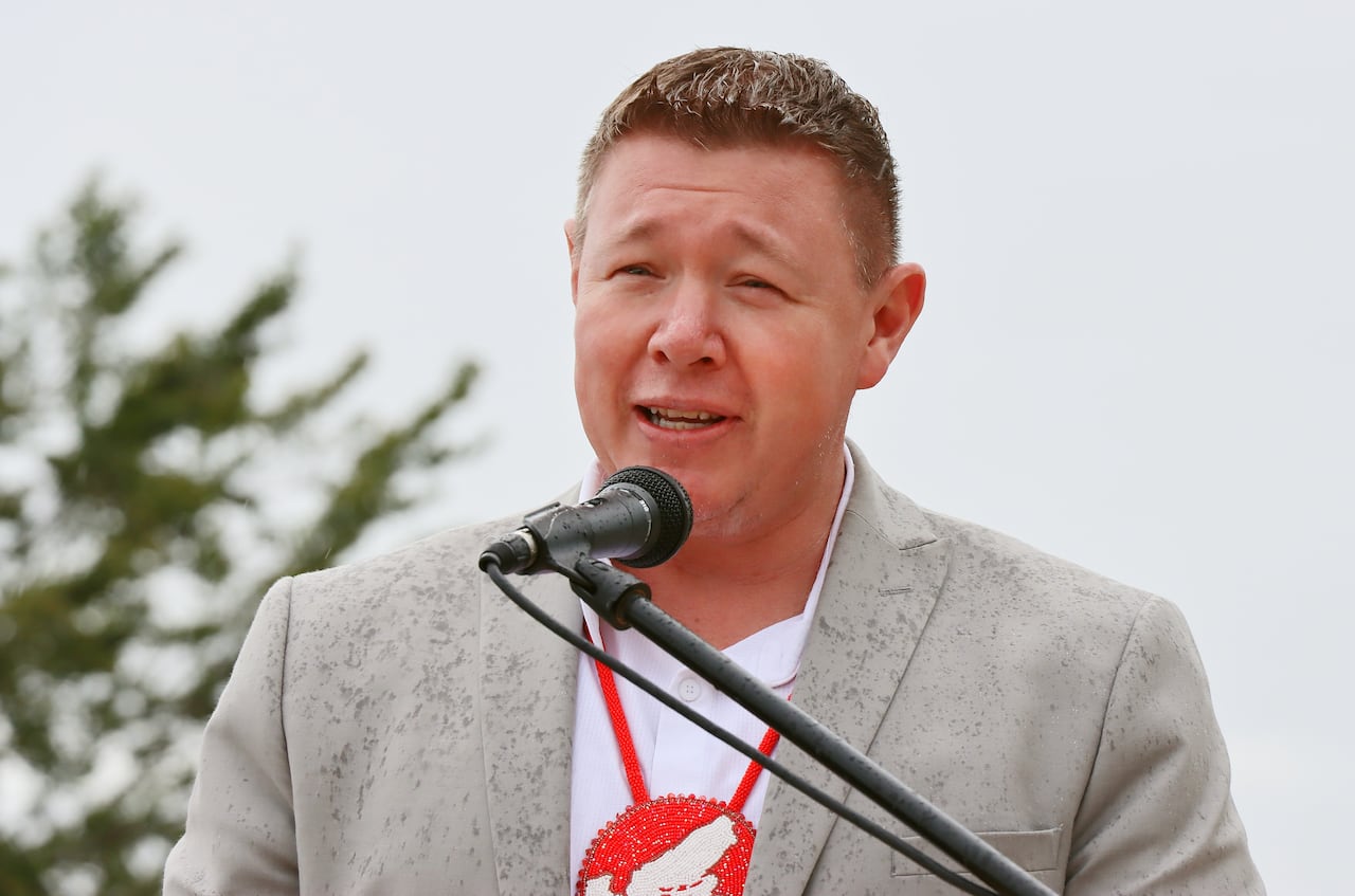 A man with short brown hair wearing a grey suit and white shirt with a red medallion around his neck speaks into a microphone.