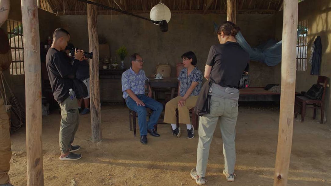 A Vietnamese father and daughter sit down in a room with a dirt floor as camera operators film their chat.