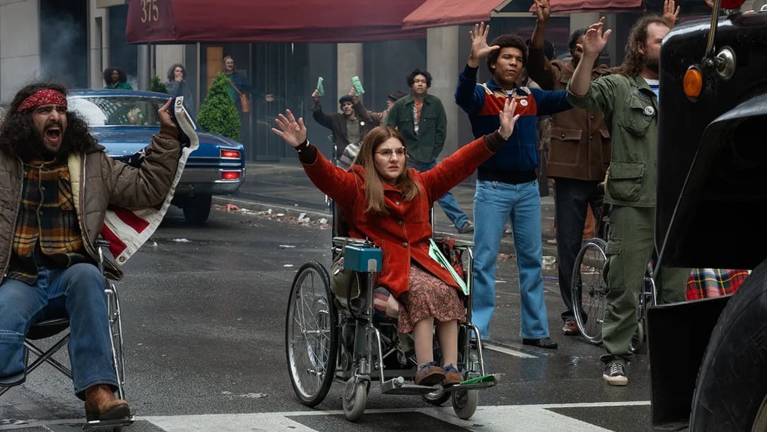 a white woman in a wheelchair sits in the middle of the street, protesting as others join in.