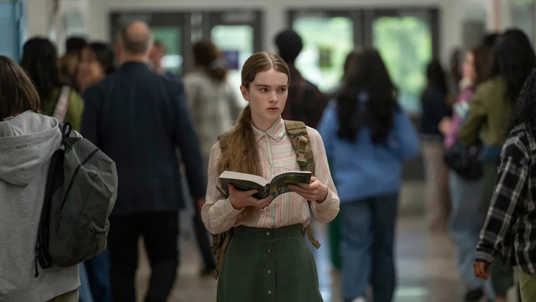 a white high school girl walks through a hallway, holding an open book.