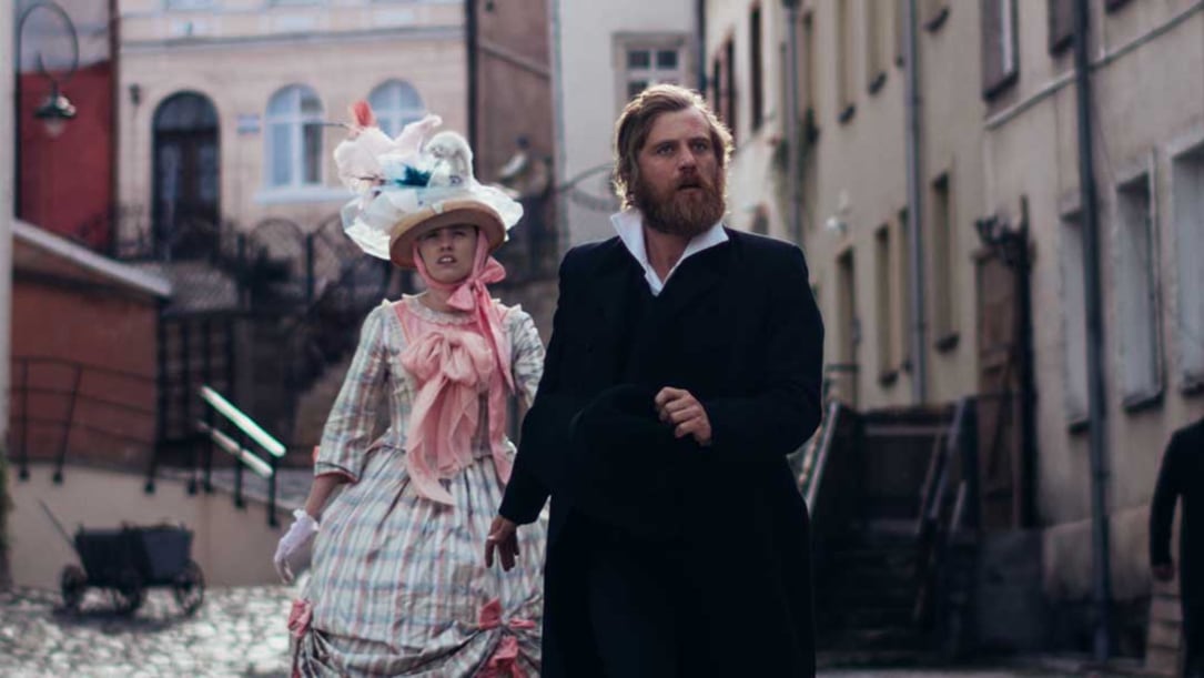 a white man and woman dressed in 1800s period garb walk down a cobblestone street.