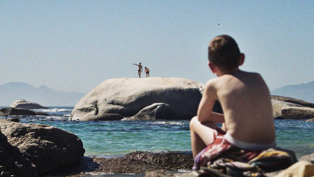 a young white boy sits on the beach watching two people standing on a rock.