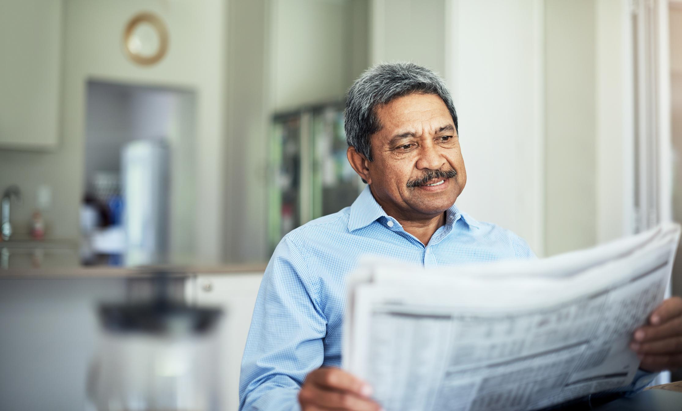 A smiling person is reading a financial newspaper while seated at a table in their home.