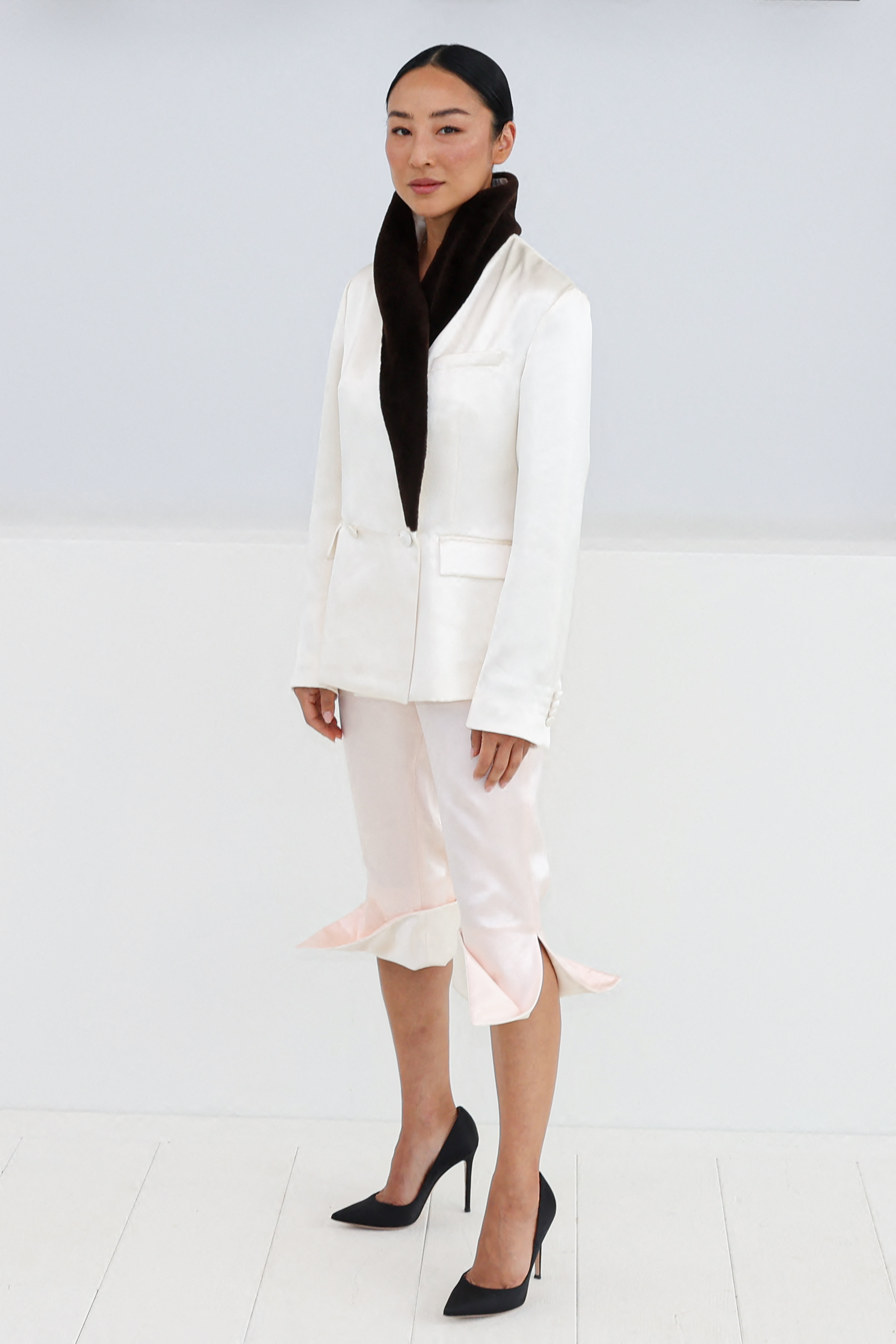 Greta Lee in a cream blazer with a dark fur scarf, light pink cropped pants, and black heels.