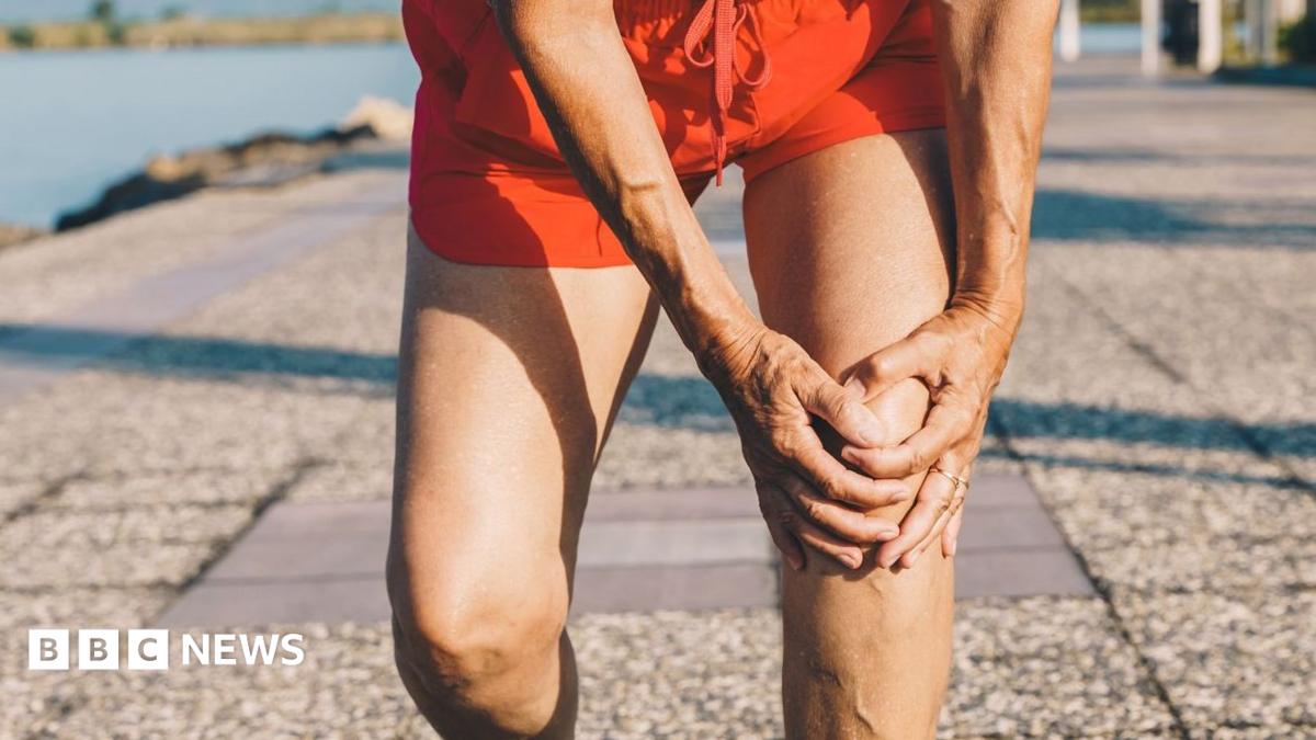 A close-up of someone holding their knee, bent over, on pavement beside a body of water.