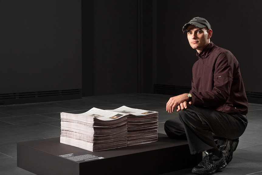 A man crouches next to a stack of newspapers.