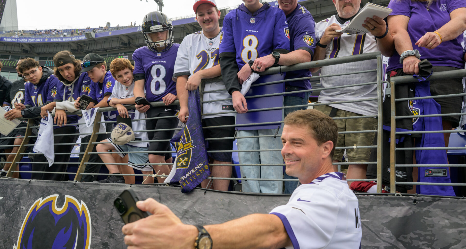 Reid Wiseman holds up a black phone to take a selfie with Baltimore Ravens fans at a football game. The fans are dressed in Ravens jerseys and shirts in purple and white. The fans are in the stands above, while Wiseman is on the field just below.