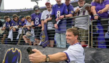 Reid Wiseman holds up a black phone to take a selfie with Baltimore Ravens fans at a football game. The fans are dressed in Ravens jerseys and shirts in purple and white. The fans are in the stands above, while Wiseman is on the field just below.
