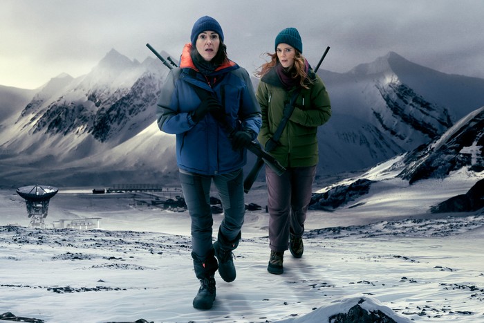 574828 Suranne Jones and Rose Leslie in Vigil, walking across a snowy landscape wearing coats, beanie hats and holding guns.