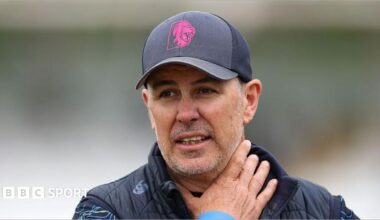 Ryan Campbell wearing a Durham gilet and navy blue cap with his right arm clutched up towards his neck