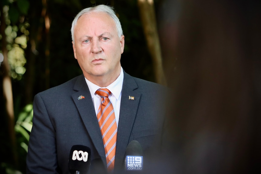 A man, NT Health Minister Steve Edgington, in a suit and orange tie stands in front of media microphones.