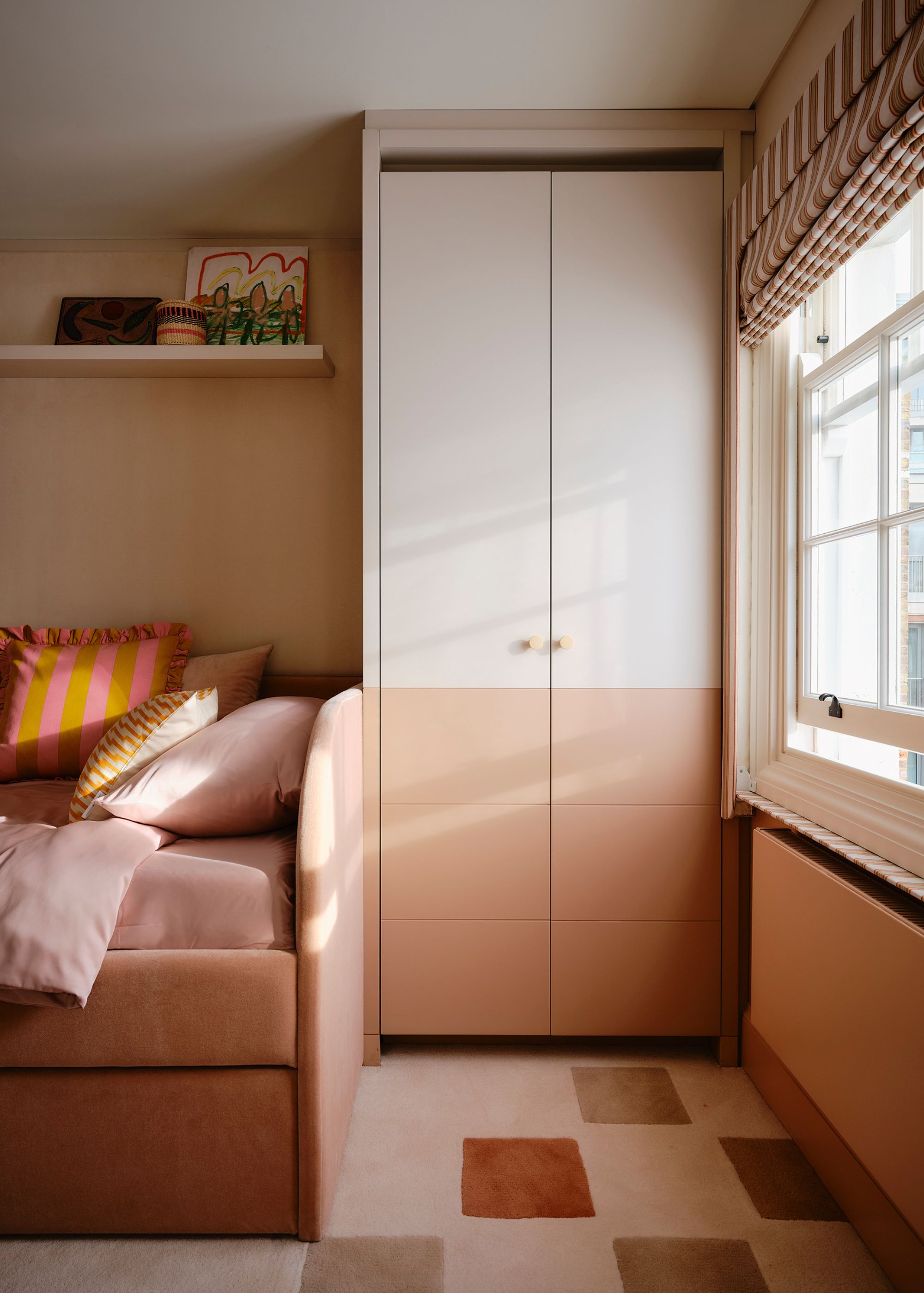 A bedroom with a built-in wardrobe painted dusky pink on the bottom half, with a soft pink neutral above. A geometric carpet in a palette of soft pink, a pink daybed with layered bedding, a gallery shelf, and striped Roman blinds.