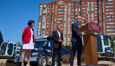 Mark Carney stands at a podium at a construction site, in front of a large, multi-storey apartment building.