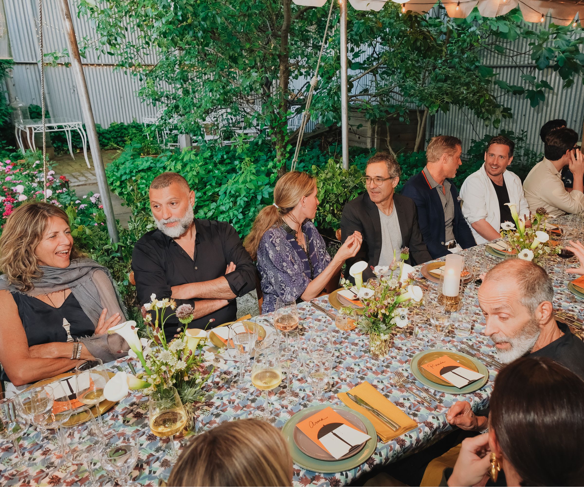 Photo of a large group of people sitting around a long rectangular dining table outdoors. Table covered in floral tableware and dinnerware, garden in the background