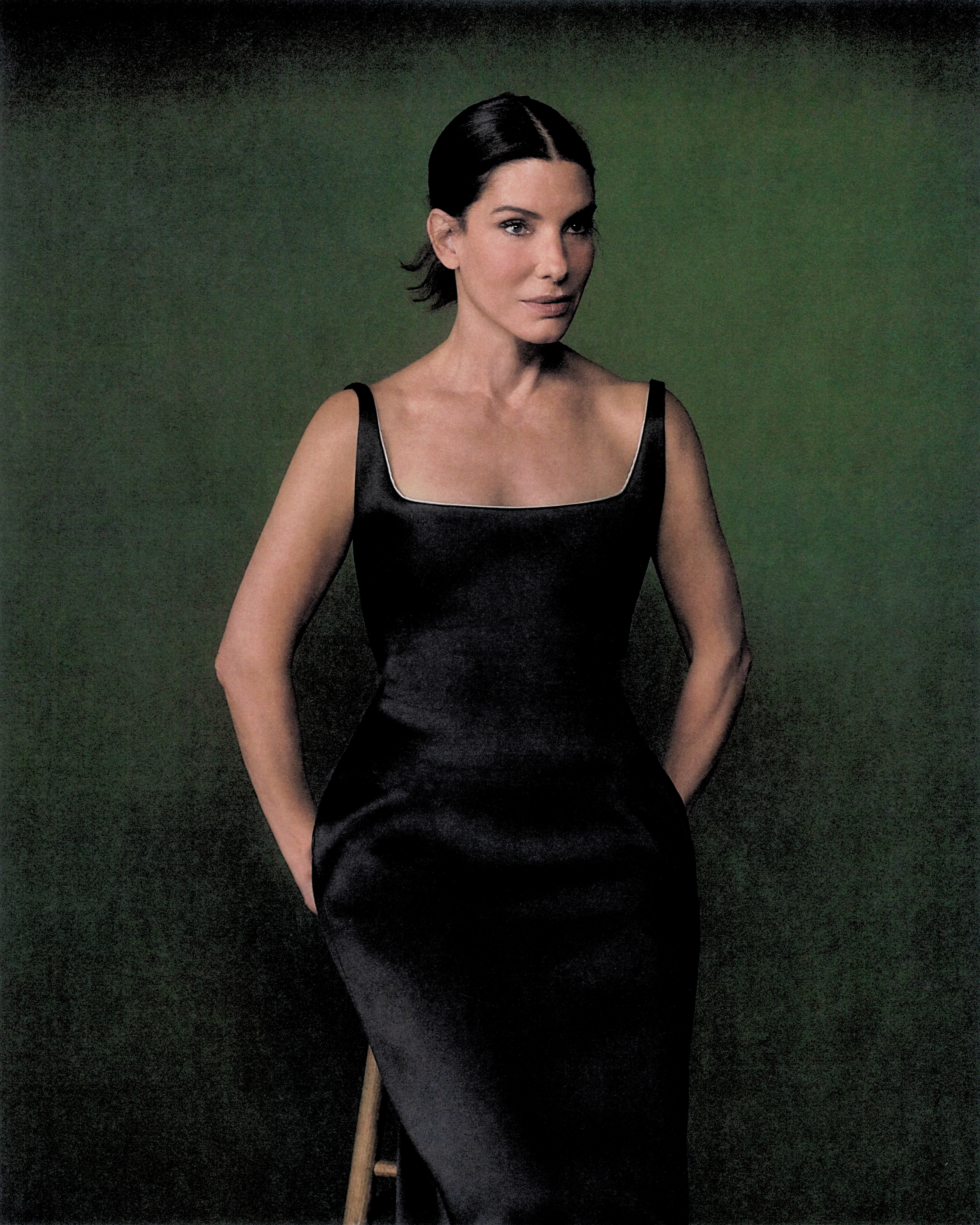 Sandra Bullock sits on a stool before a green wall with her arms behind her.
