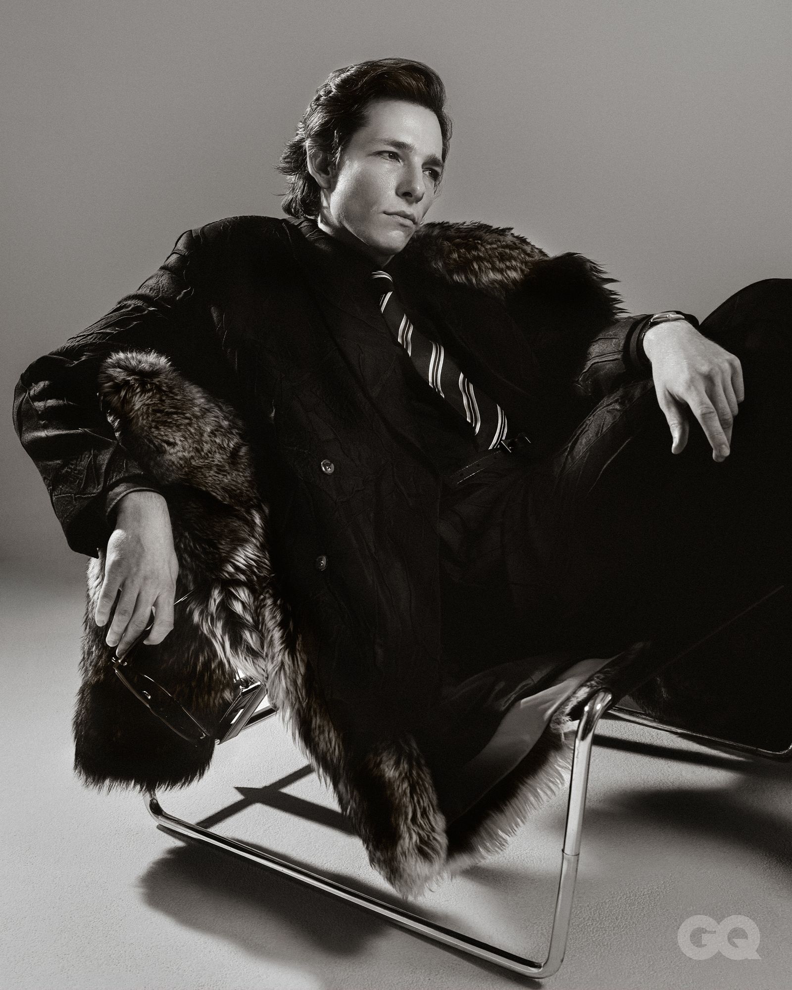 Mike Faist sitting in a chair wearing a black suit and tie