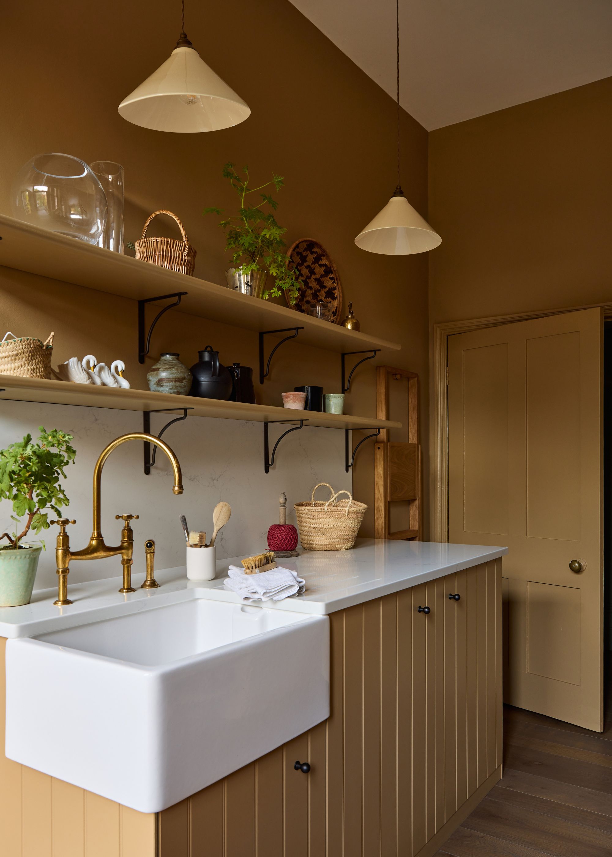 A utility room painted a caramel neutral color with matching cabinets. A Belfast sink with brass taps, open shelving with rustic decor, white counters, and two minimal pendant ceiling lights.