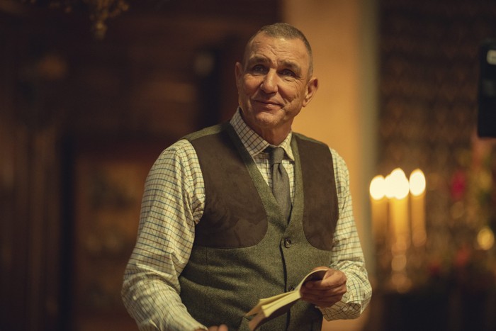 Vinnie Jones in The Gentlemen in a checkered shirt and vest holding a book, standing indoors with warm lighting, blurred background features candles and wooden decor, creating a cozy, intimate atmosphere.