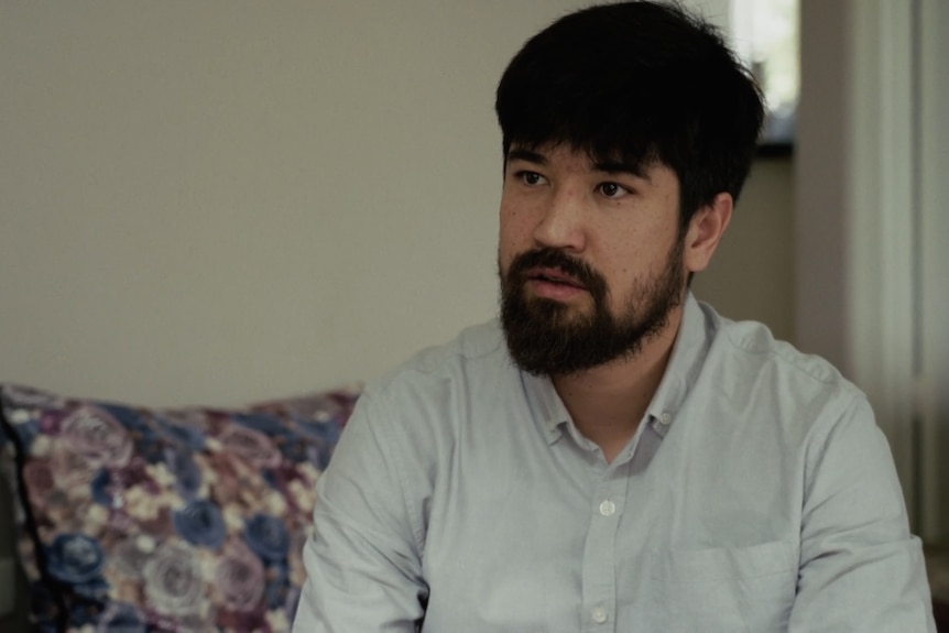 Mohammad speaks with a serious expression to someone out of view in a lounge room sat on a floral sofa.