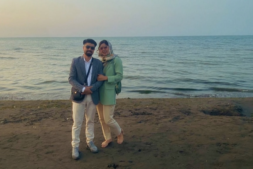 Two people pose on a beach with calm water while holding each other in a light embrace.