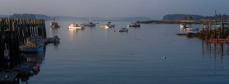 A calm harbor at dawn features several moored fishing boats, a wooden pier on the left, and distant forested islands.