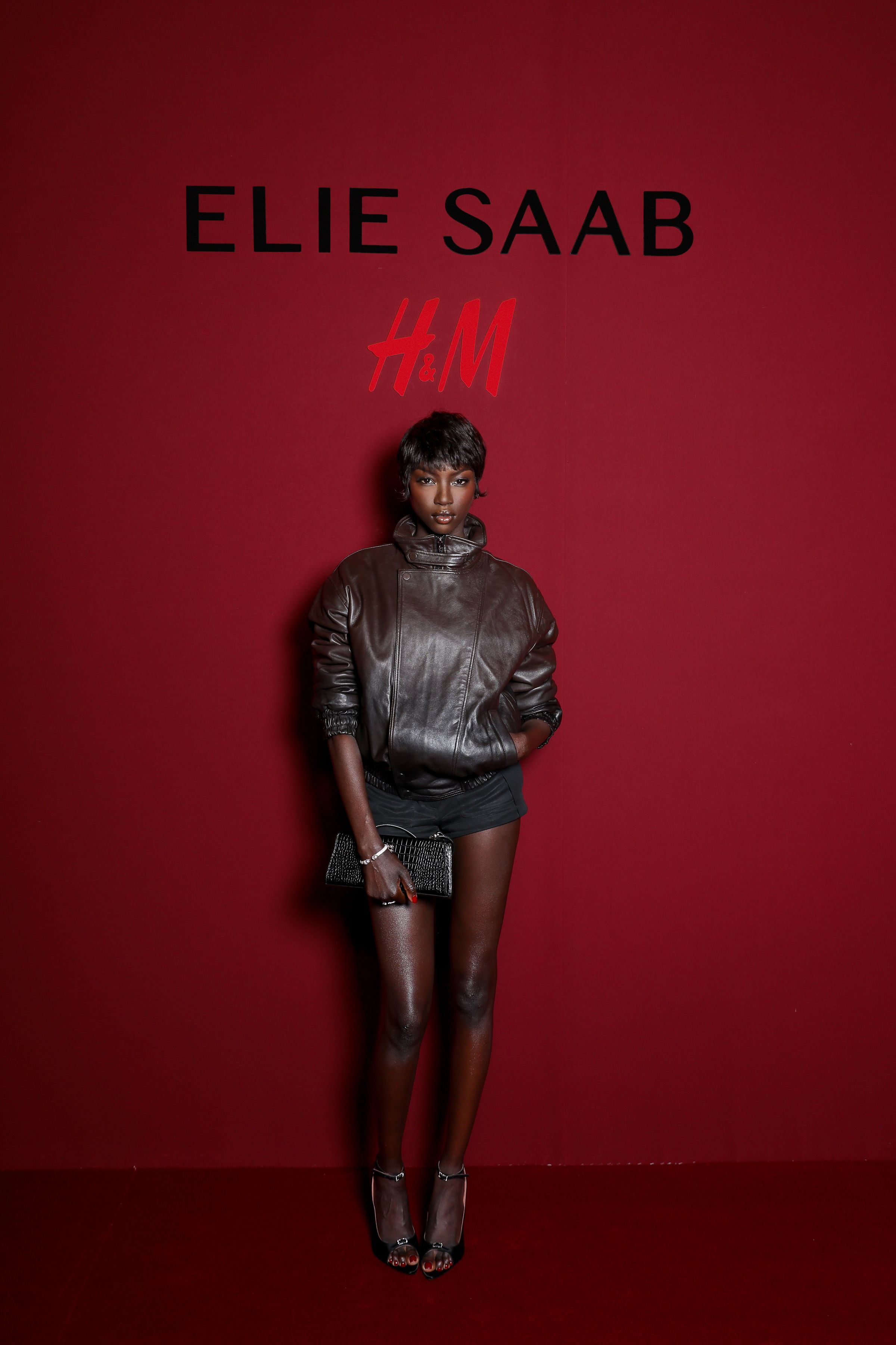 Anok Yai poses in a dark brown leather jacket and black shorts in front of an Elie Saab and H&M backdrop.