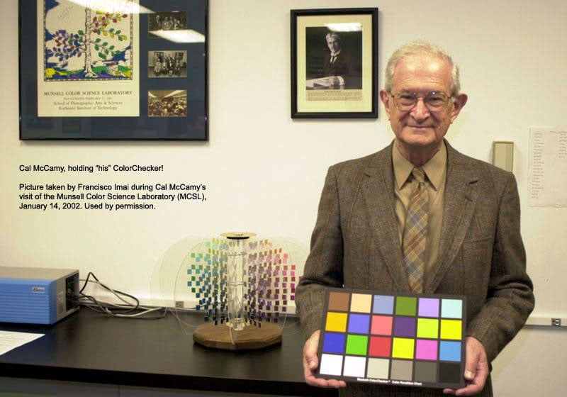 An older man in a brown suit holds a color checker chart in front of a desk with a color model display and framed pictures on the wall.