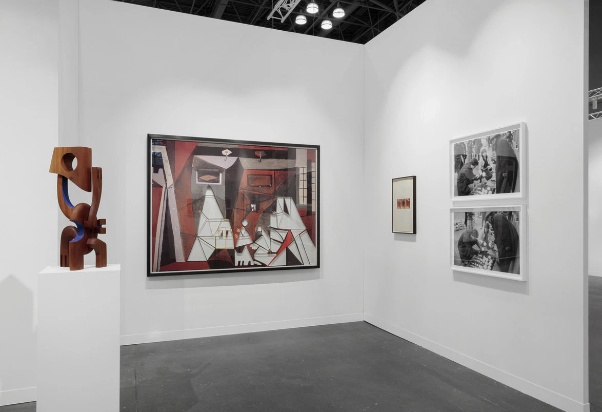 Art exhibit featuring abstract painting, wooden sculpture on pedestal, and black-and-white photographs on gallery walls.