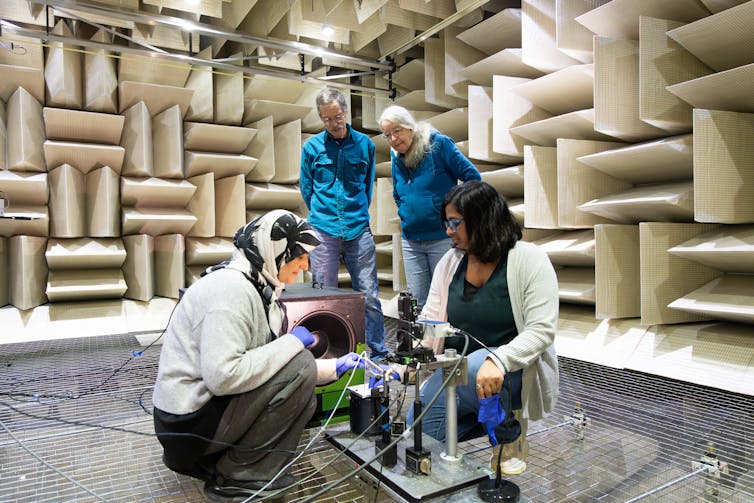 Four people, two standing and observing and two crouching on the ground next to a mechical platform, standing in a chamber with foam tiles covering the walls