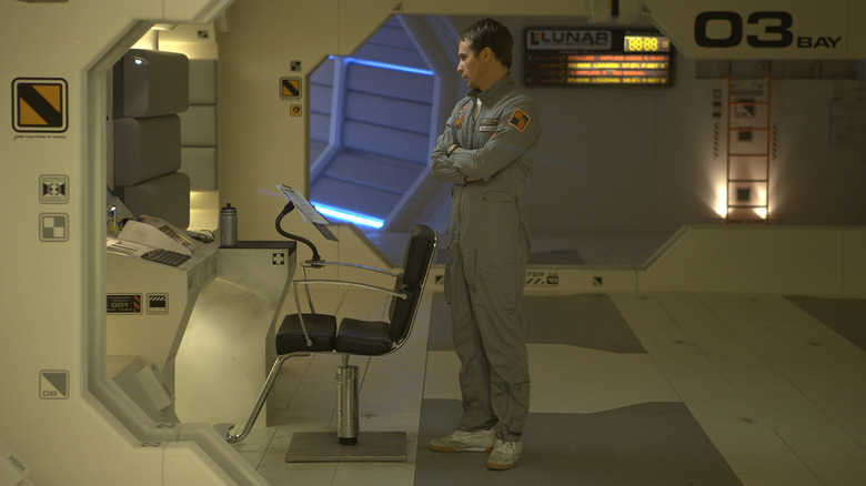 Sam Rockwell as Sam Bell, standing with his arms crossed within the clinical interior of his space station.