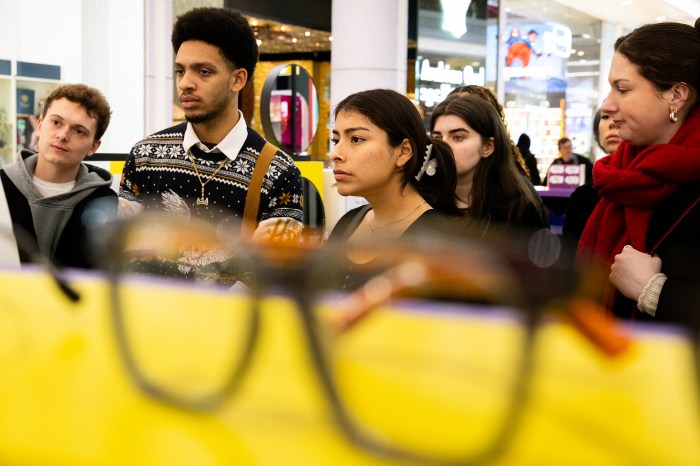 A group of students listens closely in a Pop Specs store, seen past a blurred pair of eyeglasses on a yellow display in the foreground. 