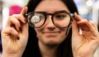 A student holds a pair of eyeglasses up to the camera, with a small metal mechanism magnified through one lens and their smiling face blurred behind the frames.