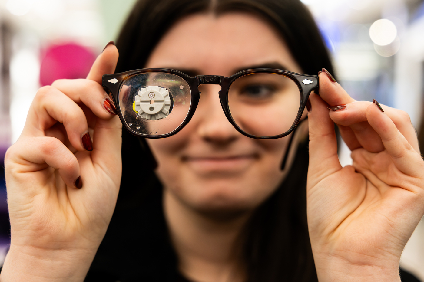 A student holds a pair of eyeglasses up to the camera, with a small metal mechanism magnified through one lens and their smiling face blurred behind the frames.