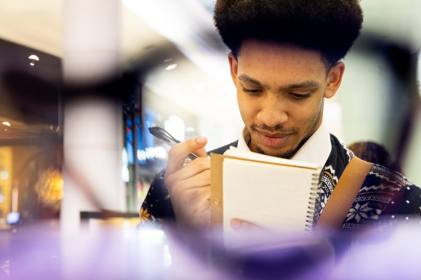 A student takes notes in a spiral notebook, framed by blurred eyeglasses in the foreground.