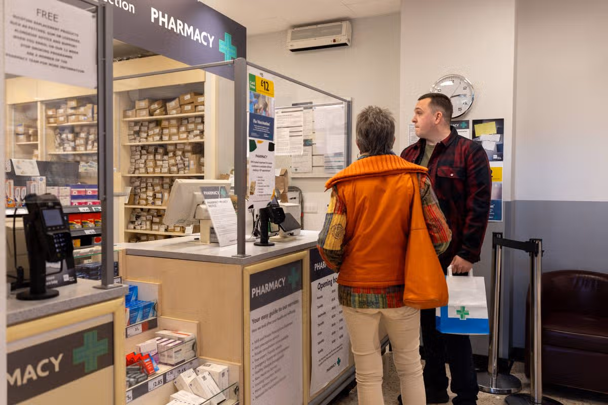 A wide shot of a grandson helping his grandmother pick up a prescription, showcasing family care and support in a pharmacy setting. They are wearing casual clothing at a shop in the North East of England.