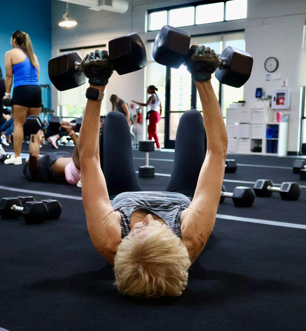 A person lifting dumbbells on a mat in a gym filled with others exercising. A person lifting dumbbells on a mat in a gym filled with others exercising.