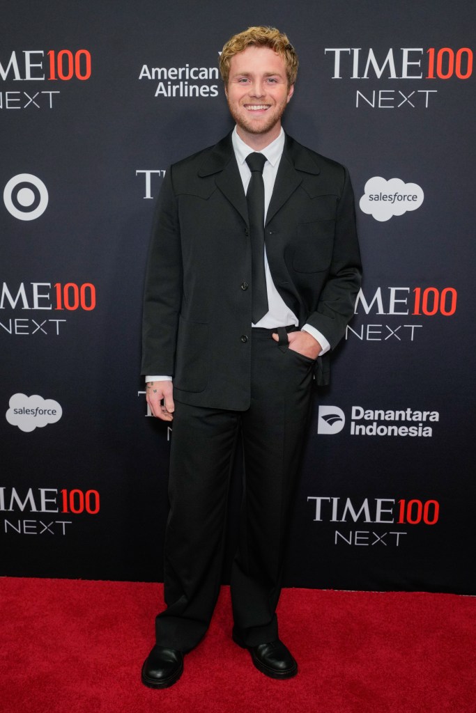 Alex Warren posing on a red carpet in a black suit and tie.