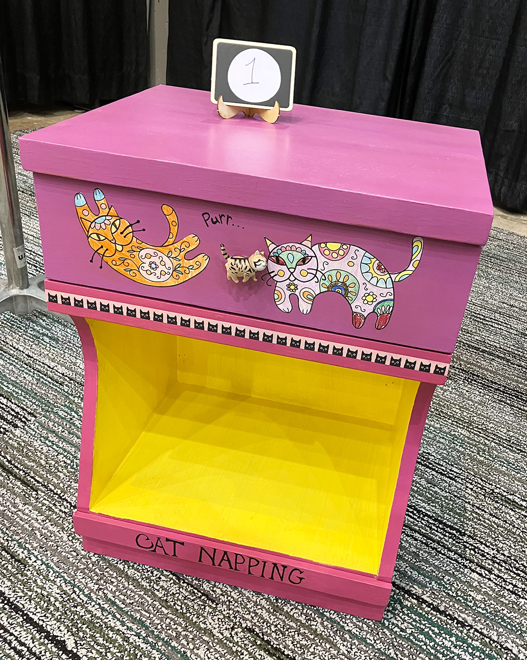 a photo of Matilda Redding's refinished nightstand. It has an open shelf (painted bright yellow) and above that a drawer with hand colored cat decals and a cute tiger drawer knob. The shelf the drawer sits on is bordered by teeny black and white cats. The rest of the nightsand is a cheerful bright pink.